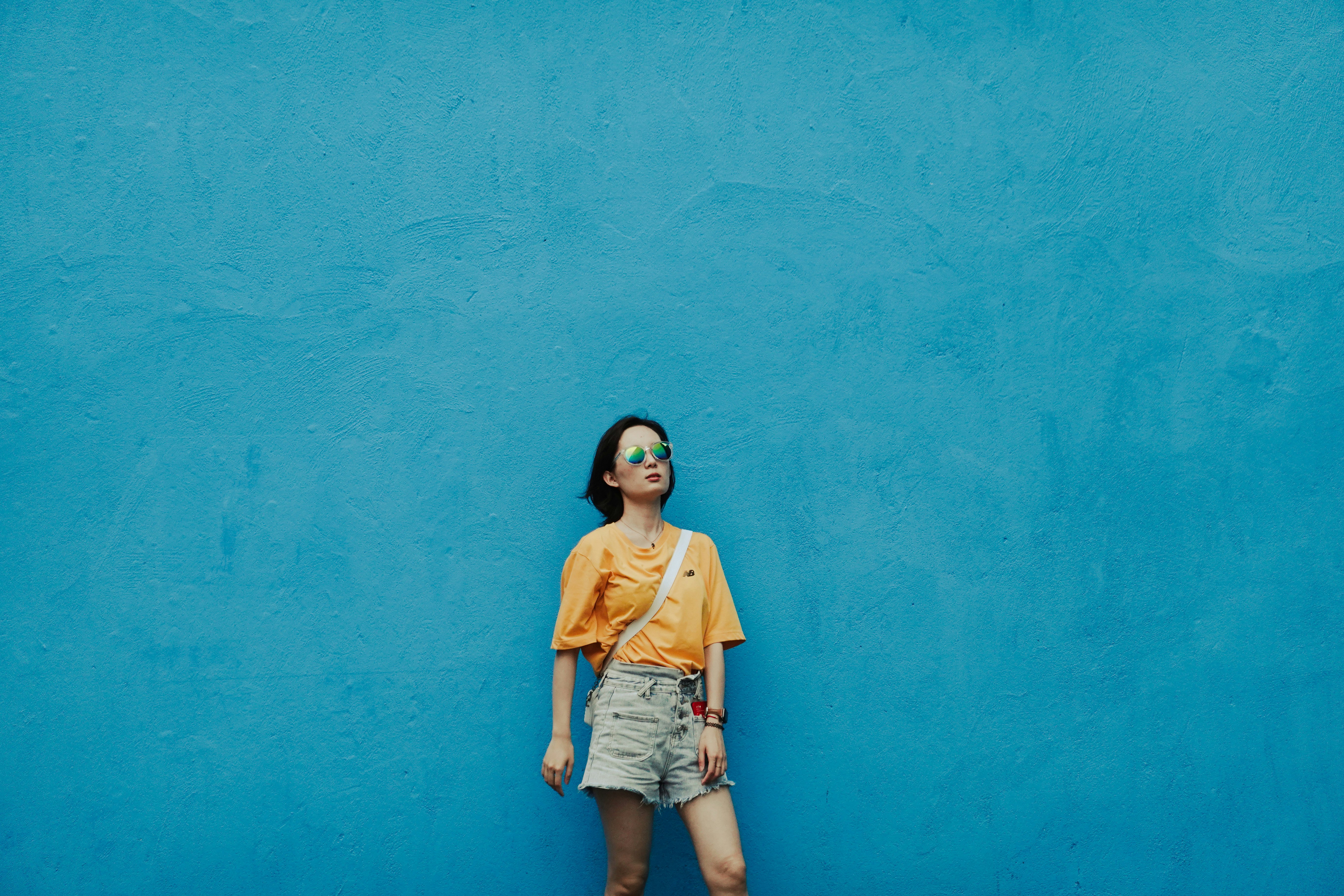 woman in yellow t-shirt and blue denim shorts standing beside blue wall